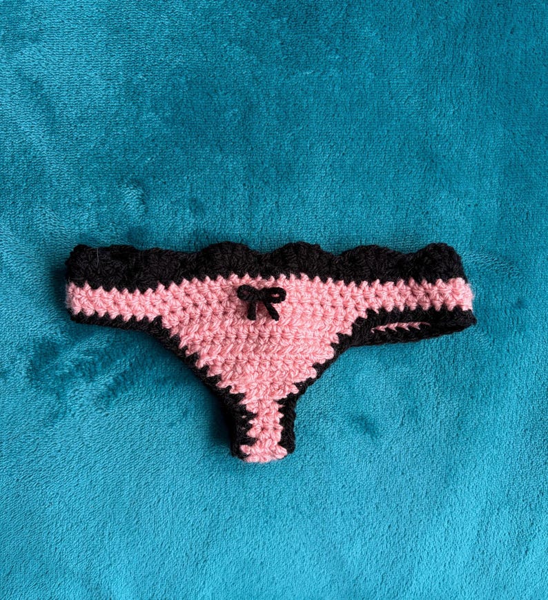 May include: A crochet thong featuring a light pink center panel with a small black bow at the center, bordered by a scalloped black trim along the waistband and leg openings, crafted with a textured yarn stitch pattern throughout the garment.