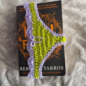 May include: A handmade crochet bookmark featuring a lime green center panel with a scalloped lavender border and a small decorative bow. The bookmark is positioned vertically over a black book cover with orange graphic details and white text.