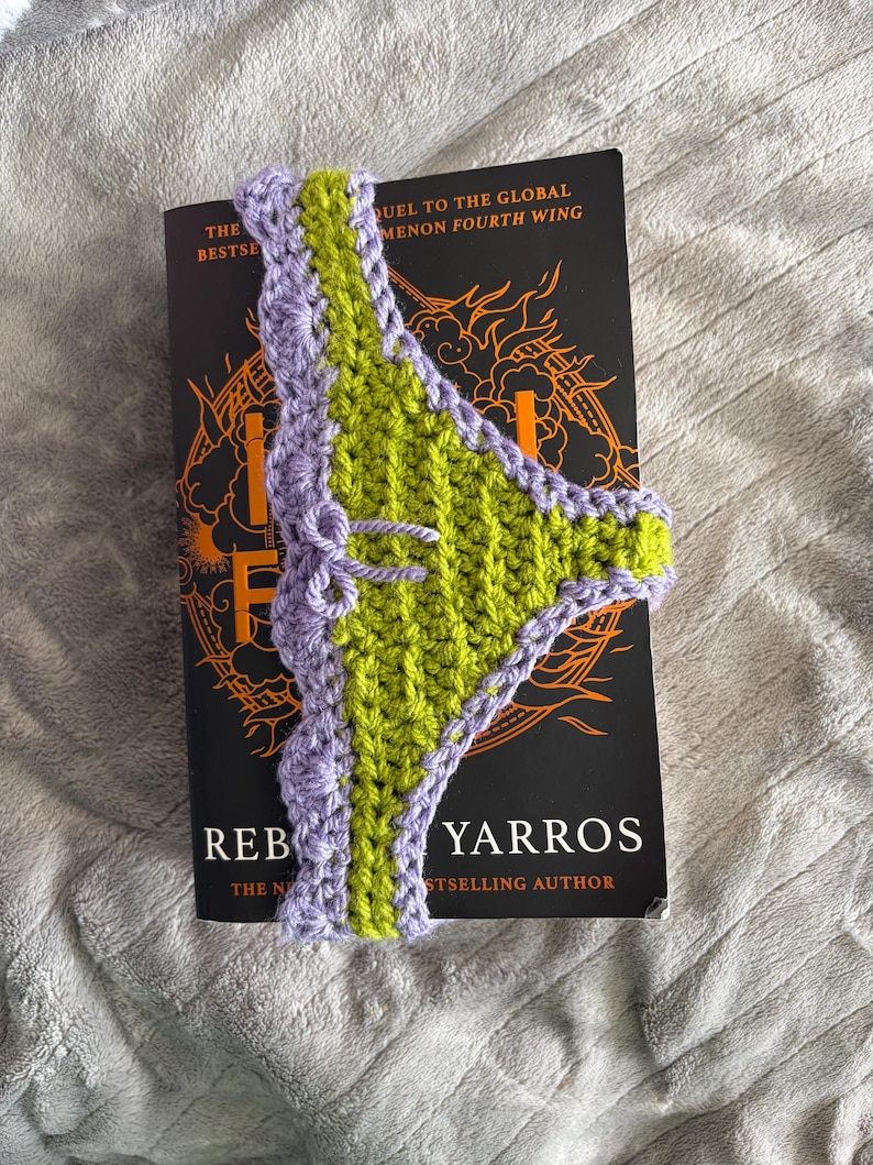 May include: A handmade crochet bookmark featuring a lime green center panel with a scalloped lavender border and a small decorative bow. The bookmark is positioned vertically over a black book cover with orange graphic details and white text.
