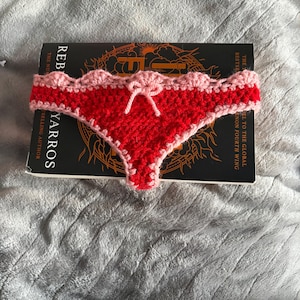 May include: A handmade crochet bikini bottom in bright red yarn with a light pink scalloped trim along the waistband and leg openings. The center front features a small, decorative light pink crochet bow tied in a knot.