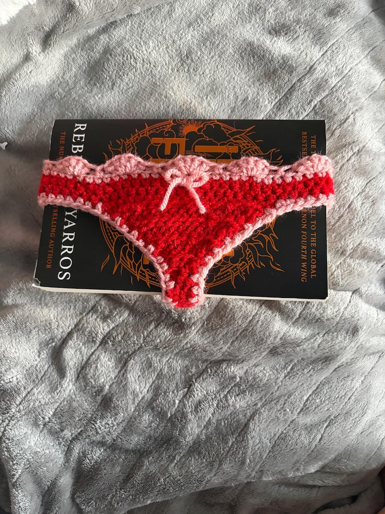 May include: A handmade crochet bikini bottom in bright red yarn with a light pink scalloped trim along the waistband and leg openings. The center front features a small, decorative light pink crochet bow tied in a knot.
