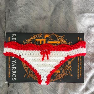 May include: A crochet bikini bottom featuring a white triangular center panel with horizontal ridges, bordered by a scalloped red trim along the top and sides. A small red yarn bow is centered at the top edge of the white section.
