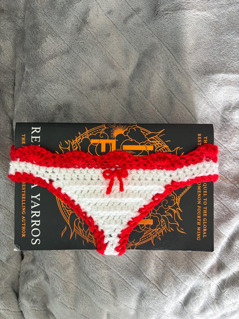 May include: A crochet bikini bottom featuring a white triangular center panel with horizontal ridges, bordered by a scalloped red trim along the top and sides. A small red yarn bow is centered at the top edge of the white section.