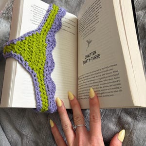 May include: A crochet bookmark featuring a central lime green vertical strip bordered by a scalloped lavender edge. The bookmark is placed inside an open book, with the text Chapter Forty-Three visible on the right page, showing a dragon icon.