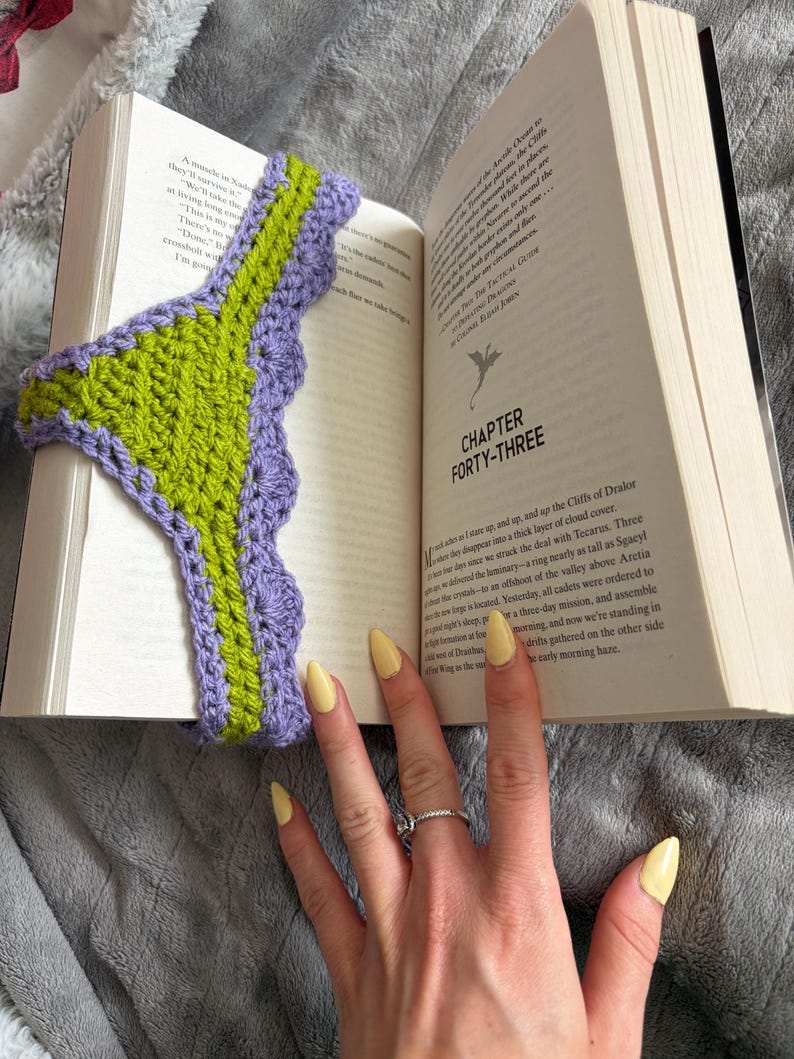 May include: A crochet bookmark featuring a central lime green vertical strip bordered by a scalloped lavender edge. The bookmark is placed inside an open book, with the text Chapter Forty-Three visible on the right page, showing a dragon icon.
