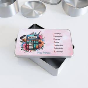 Può includere: Una scatola di metallo rettangolare con un coperchio con un design colorato. Il design include una calcolatrice, una tazza di caffè e una composizione floreale. Il testo sul coperchio recita "TEACHER LIFE" e un elenco di attributi positivi. La scatola è aperta, rivelando l'interno.