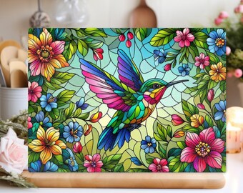 Pode incluir: Uma tábua de corte retangular de vidro com uma ilustração colorida em estilo vitral de um beija-flor em voo rodeado por flores e folhas verdes. O design utiliza contornos pretos espessos para definir as formas florais e da ave.