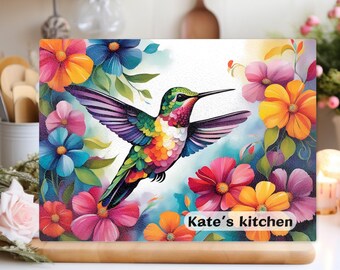 Pode incluir: Tábua de corte retangular com um beija-flor aquarela em voo, rodeado por flores coloridas. O design inclui tons de rosa, laranja, azul e verde. O texto "Kate's kitchen" é exibido no canto inferior direito.