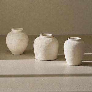 May include: Three off-white ceramic vases with textured surfaces. The vases have rounded bodies and varying heights, with a small opening at the top. The neutral color and simple design make them suitable for various home decor styles.