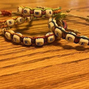 May include: Three handmade bracelets crafted from colorful braided cord and light-colored wooden beads. Each bead features a single black letter, spelling out words including IOWA, WOST, and PEACE. The bracelets have adjustable knotted cord closures.