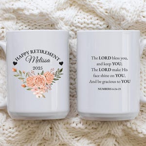 Może przedstawiać: Biały ceramiczny kubek z kwiatowym wzorem i napisem "HAPPY RETIREMENT Melissa 2025". Druga strona kubka zawiera cytat religijny z Księgi Liczb 6:24-25. Kubek jest umieszczony na białym, dzianinowym tle.