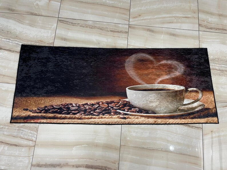Puede incluir: Una alfombra de bienvenida marr&oacute;n y negra con una taza de caf&eacute; y granos de caf&eacute;. La taza de caf&eacute; es blanca con un borde marr&oacute;n y un vapor en forma de coraz&oacute;n que sale de ella. Los granos de caf&eacute; est&aacute;n esparcidos alrededor de la taza.