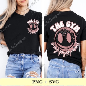 Puede incluir: Camiseta negra con un diseño de cara sonriente rosa y el texto "THE GYM & my psych ward". El diseño está impreso en la parte delantera y trasera. La camiseta se combina con vaqueros azules.