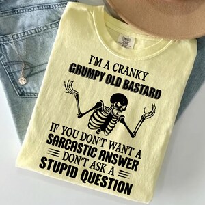 Könnte beinhalten: Ein blassgelbes T-Shirt mit einer schwarzen Grafik eines Skeletts mit erhobenen Armen. Der Text lautet: I'M A CRANKY GRUMPY OLD BASTARD IF YOU DON'T WANT A SARCASTIC ANSWER DON'T ASK A STUPID QUESTION. Das T-Shirt hat einen Rundhalsausschnitt und ein Etikett.