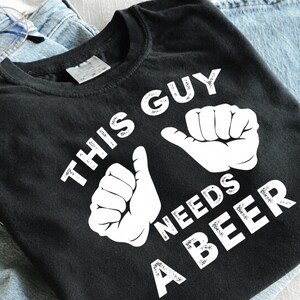 May include: A black short-sleeved t-shirt featuring white graphic text that reads THIS GUY NEEDS A BEER, accompanied by two white illustrations of hands with thumbs pointing upward, positioned centrally on the front of the garment.