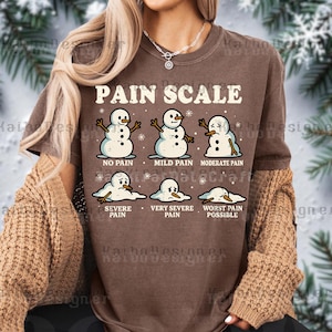 May include: Brown t-shirt with a graphic of a pain scale using snowmen. The text reads "PAIN SCALE" above the snowmen, with pain levels from "No Pain" to "Worst Pain Possible".