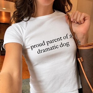 May include: A white short-sleeved t-shirt featuring the black text "proud parent of a dramatic dog" printed across the chest. The garment has a crew neckline and a fitted silhouette, paired with a brown leather shoulder bag with gold hardware.