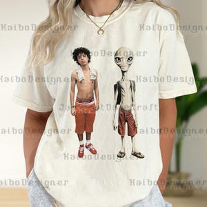 May include: Cream-colored t-shirt featuring a graphic of two figures: a person in orange shorts and red sneakers, and an alien in red shorts and sandals. The shirt has the text "Kaibo Designer" printed on it.