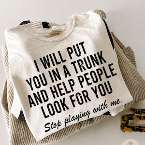 May include: A cream-colored t-shirt featuring bold black text that reads: I WILL PUT YOU IN A TRUNK AND HELP PEOPLE LOOK FOR YOU Stop playing with me. The shirt is layered over a beige knit garment, with a small tortoiseshell hair clip visible nearby.