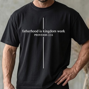 May include: Black t-shirt with the text "fatherhood is kingdom work" and "PROVERBS 22:6" in white. A vertical white line is centered above the text. The shirt is a crew neck and has short sleeves.