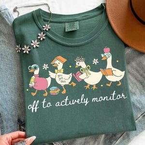 May include: Dark green t-shirt featuring a whimsical illustration of four cartoon geese, each with unique accessories like glasses and hats, carrying books. The text "off to actively monitor" is printed below the geese. A floral necklace and a brown hat are also visible.