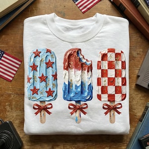 May include: White sweatshirt featuring three popsicle designs in red, white, and blue. One popsicle is blue with red stars, another is red and white with a bite taken out, and the last is a red and white checkerboard pattern. Each popsicle has a wooden stick and a red bow.