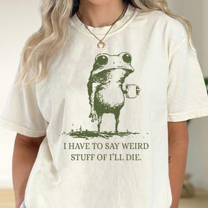 Op de afbeelding: Crèmekleurig T-shirt met een groene illustratie van een kikker die een mok vasthoudt. De kikker heeft grote ogen en een enigszins chagrijnige uitdrukking. De tekst eronder luidt: "I HAVE TO SAY WEIRD STUFF OF I'LL DIE."
