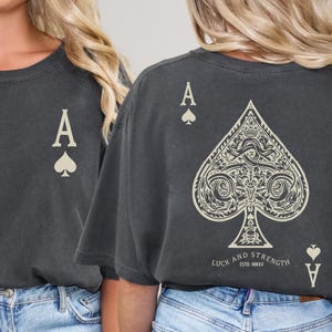 Op de afbeelding: Donkergrijs t-shirt met een ontwerp van een schoppen aas kaart. De voorkant van het shirt heeft een kleine schoppen aas afbeelding, terwijl de achterkant een groot, gedetailleerd schoppen ontwerp toont met de tekst "LUCK AND STRENGTH ESTO.MMXV".