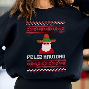 Può includere: Felpa nera con un design festivo. La grafica presenta Babbo Natale con un sombrero e occhiali da sole, con la scritta "FELIZ NAVIDAD" in bianco. Il design include motivi rossi, verdi e bianchi.