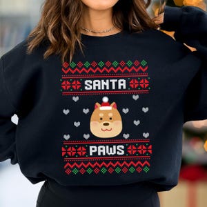 Può includere: Felpa nera con un design festivo. Il design include le parole bianche "SANTA PAWS", un cane Shiba Inu con un cappello da Babbo Natale e motivi geometrici rossi e verdi. Cuori bianchi sparsi.