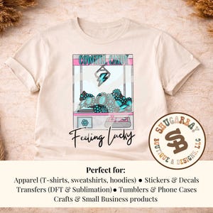 Puede incluir: Camiseta color crema con un dise&ntilde;o gr&aacute;fico de una m&aacute;quina de garras con el texto "COWGIRL CANDY" y "Feeling Lucky". La m&aacute;quina de garras contiene objetos turquesa y plateados. La camiseta es de Shugarbay Boutique & Designs.