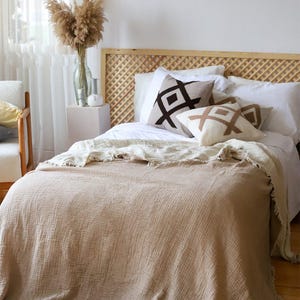 Può includere: Un letto con una coperta strutturata marrone chiaro e cuscini decorativi con disegni geometrici. La testiera è in legno intrecciato. Un vaso di fiori secchi è su un comodino.