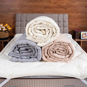 May include: Three rolled-up blankets in cream, gray, and beige rest on a bed with a white quilted mattress cover. The bed has a neutral-toned headboard and is set against a wood-paneled wall. A small table with flowers is visible.