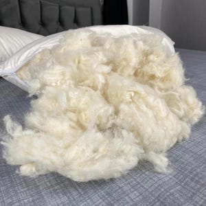 May include: A white pillow with an open zip, spilling out a large quantity of fluffy, cream-coloured filling. The filling looks loose and soft, suggesting a comfortable pillow. The pillow is on a grey bed.