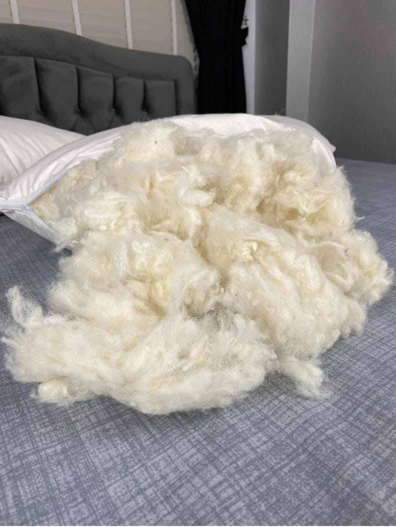 May include: A white pillow with an open zip, spilling out a large quantity of fluffy, cream-coloured filling. The filling looks loose and soft, suggesting a comfortable pillow. The pillow is on a grey bed.