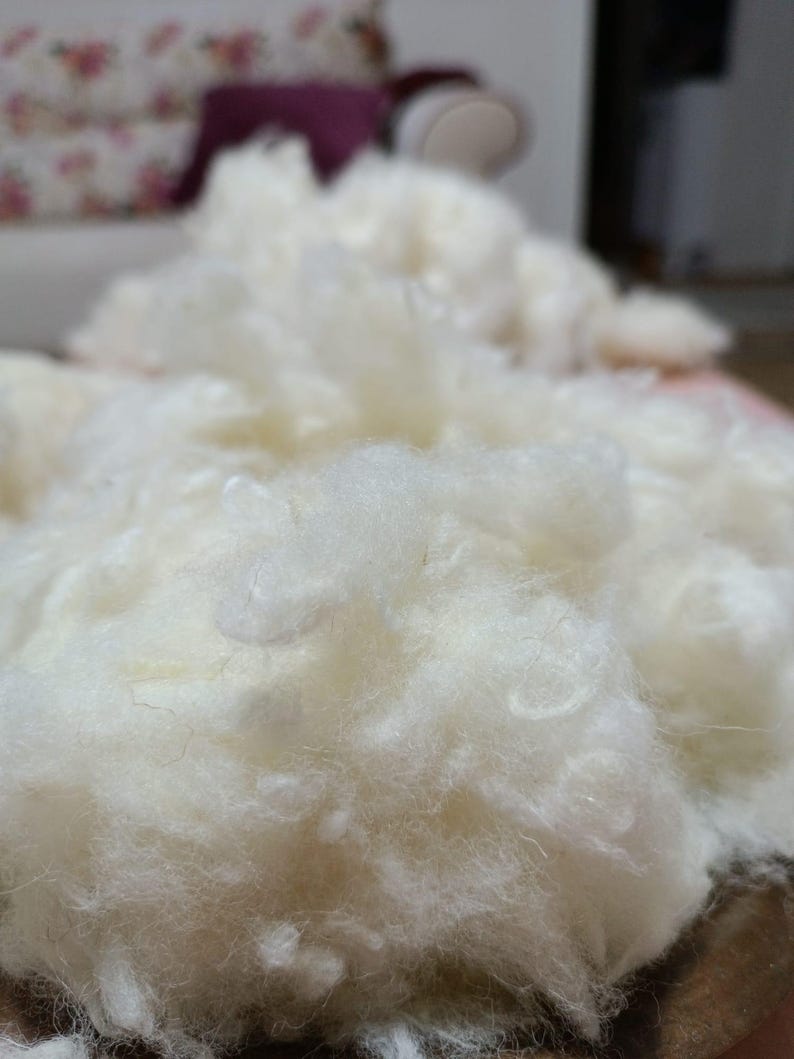 May include: Close-up of a pile of fluffy, off-white material, possibly batting or stuffing. The texture appears soft and cloud-like, with visible fibres. The background is blurred, suggesting a focus on the material's texture and volume. The material is likely used for crafts or filling.