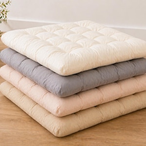 May include: A stack of four square cushions in varying neutral colors: cream, gray, light pink, and beige. Each cushion has a quilted pattern. The cushions are stacked on top of each other.