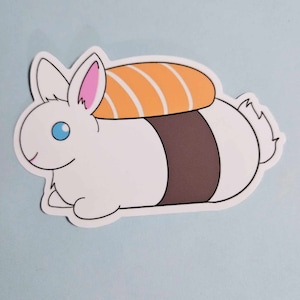 May include: A white cartoon bunny sticker with blue eyes and pink inner ears, topped with a sushi roll. The sushi roll has orange and white stripes. The bunny has a brown stripe around its middle.
