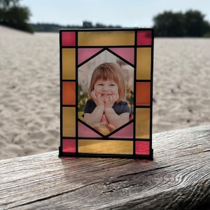Può includere: Una cornice per foto in vetro colorato con un design giallo, rosa e arancione. La cornice contiene una foto di una bambina sorridente.