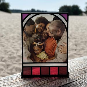 Pode incluir: Um porta-retratos de vidro colorido com um retrato de família dentro. A moldura tem uma borda roxa e branca com detalhes vermelhos. A família está sorrindo e se abraçando.