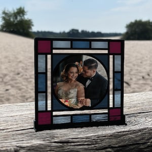 Pode incluir: Um porta-retratos de vidro colorido com uma moldura preta e painéis de vidro azul e rosa. A moldura contém uma foto de um casal sorrindo um para o outro.