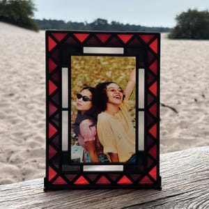 Pode incluir: Um porta-retratos de vidro vermelho com uma foto de duas mulheres usando óculos de sol. A moldura tem um padrão geométrico com detalhes vermelhos, brancos e pretos.