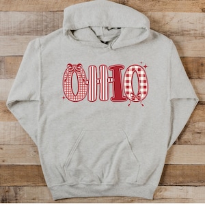 May include: A light gray hoodie with a front pocket and a hood. The word "OHIO" is printed across the chest in red. The letters are decorated with patterns, including gingham and stripes, and bows. The hoodie is made of a soft material.