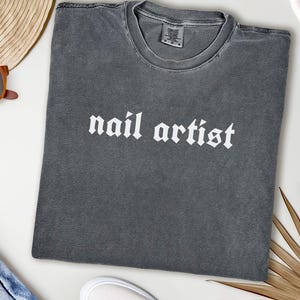 May include: Dark gray t-shirt with the words "nail artist" in white gothic font. The shirt is a Comfort Colors brand and appears to be pre-shrunk. The shirt is laid flat on a white surface, with a straw hat and sunglasses in the background.