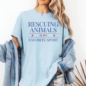 May include: Light blue t-shirt with the text "Rescuing Animals Is My Favorite Sport" in navy blue. The text is centered and includes paw print graphics. The shirt is made of a soft material and has short sleeves.