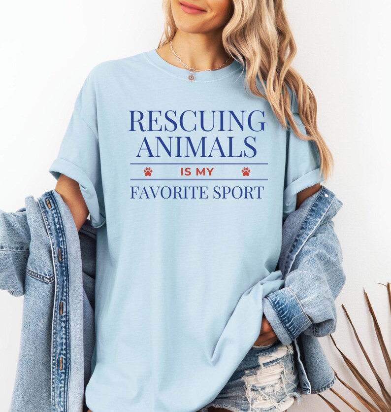 May include: Light blue t-shirt with the text "Rescuing Animals Is My Favorite Sport" in navy blue. The text is centered and includes paw print graphics. The shirt is made of a soft material and has short sleeves.