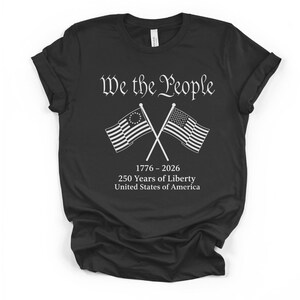 May include: Black t-shirt with the text "We the People" in a decorative font. Below the text are two crossed flags, one with a circle of stars and the other with stripes. The shirt also includes the text "1776-2026 250 Years of Liberty United States of America."
