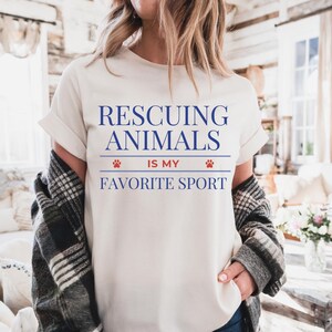 May include: Cream-colored t-shirt with the text "Rescuing Animals Is My Favorite Sport" in blue. The text is centered and includes paw print graphics. The shirt has rolled-up sleeves and is worn with a plaid jacket.