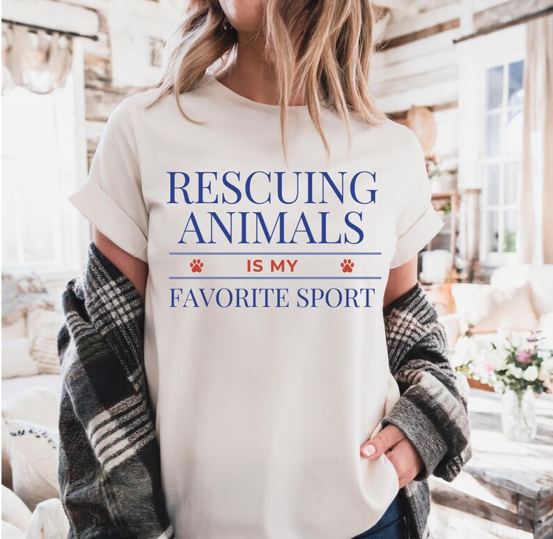 May include: Cream-colored t-shirt with the text "Rescuing Animals Is My Favorite Sport" in blue. The text is centered and includes paw print graphics. The shirt has rolled-up sleeves and is worn with a plaid jacket.