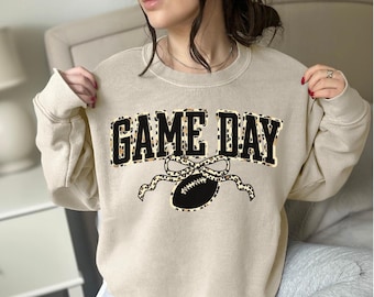 Può includere: Felpa beige con la scritta "GAME DAY" in un design a stampa leopardata nera e beige. Sotto il testo, un pallone da football americano nero con un design a fiocco e nastro leopardato.